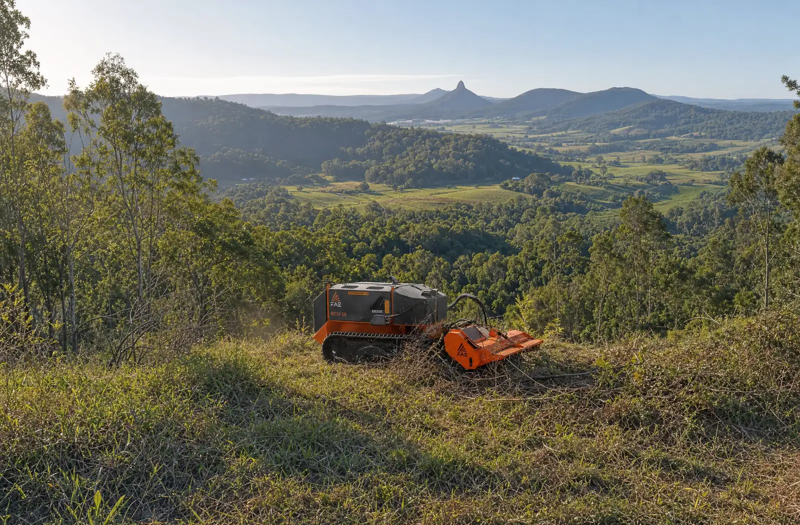 A remote-controlled tracked forestry mulcher clearing regrowth on a grassy slope above a green valley