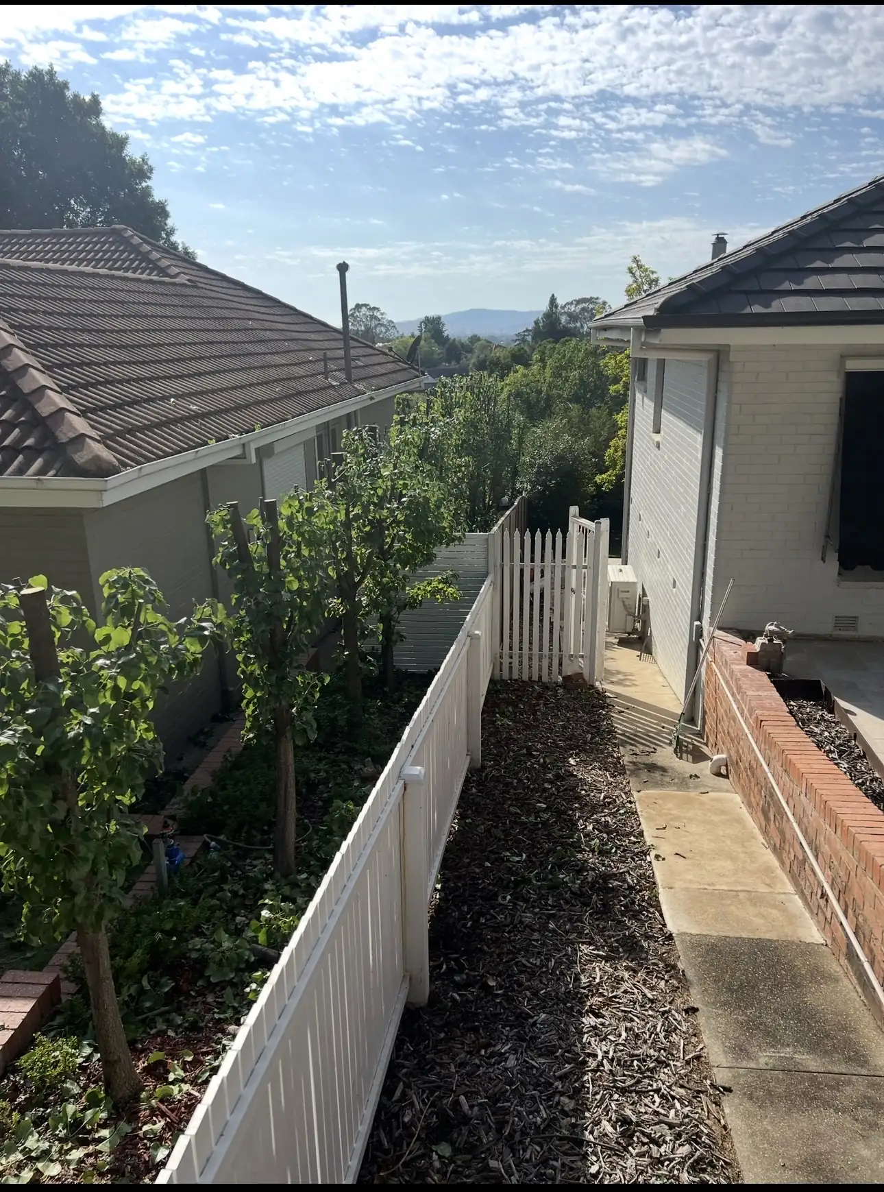 A narrow mulched path between a white fence and garden planting beside a house